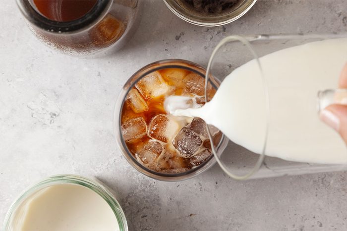 A hand pours milk into a glass of iced coffee, surrounded by jars and bowls on a light-colored surface.