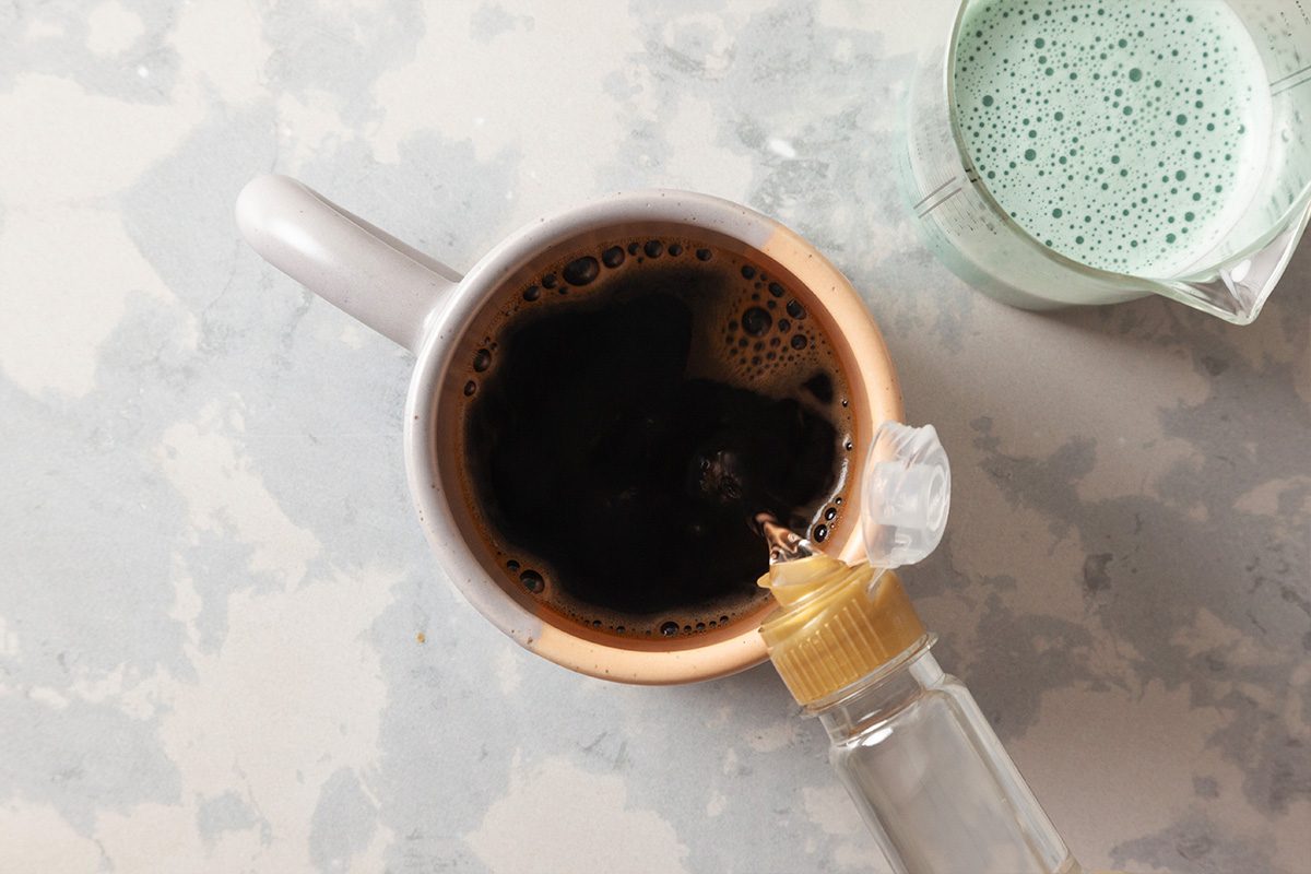 Overhead view of a mug filled with black coffee, with a bottle pouring liquid into it. Next to the mug is a small glass container holding a green frothy beverage. All items are on a light mottled surface.