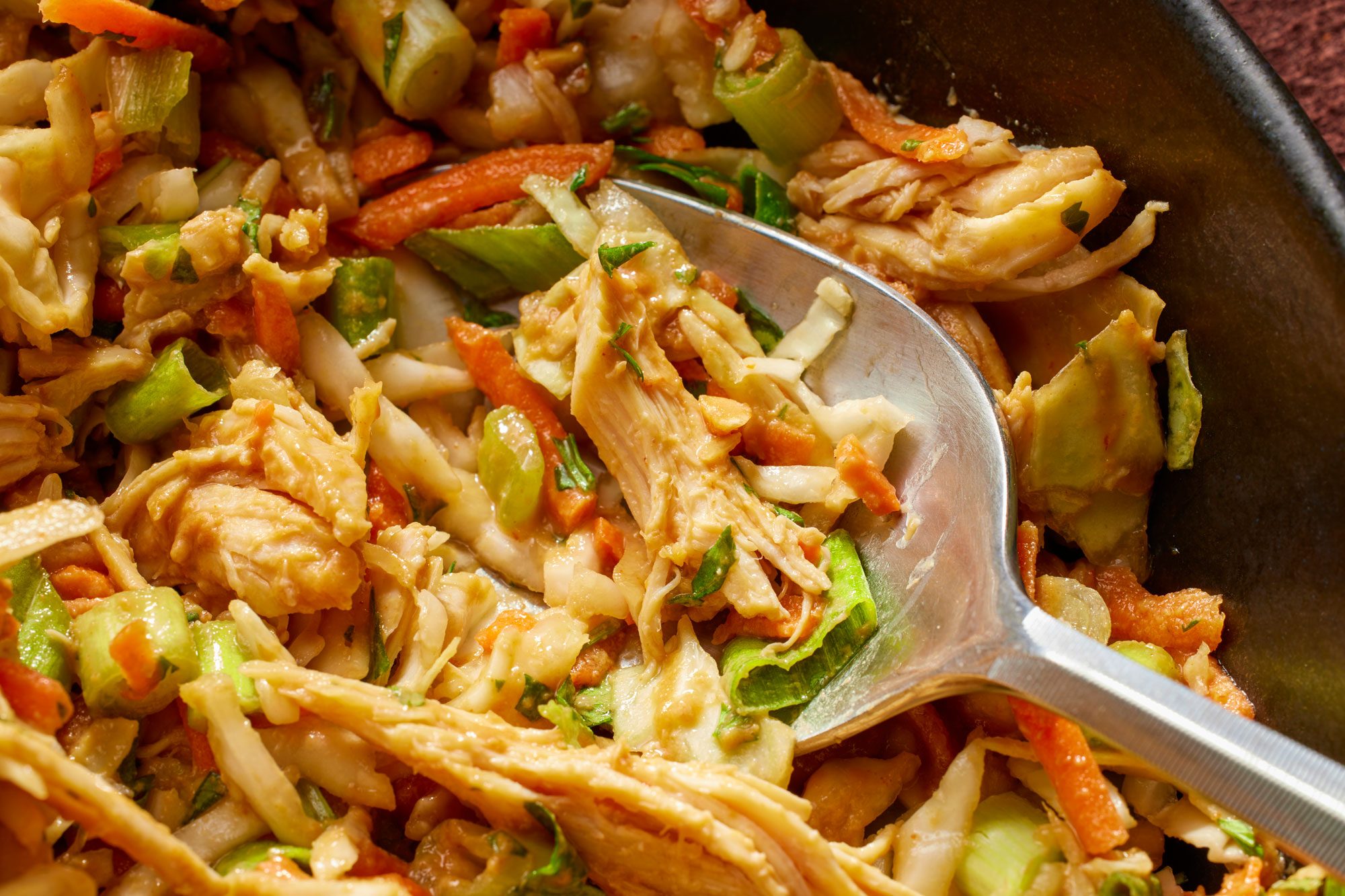 Close-up shot of Thai Chicken Coleslaw in a large bowl with cabbage, carrots, and green onions, with a silver spoon;