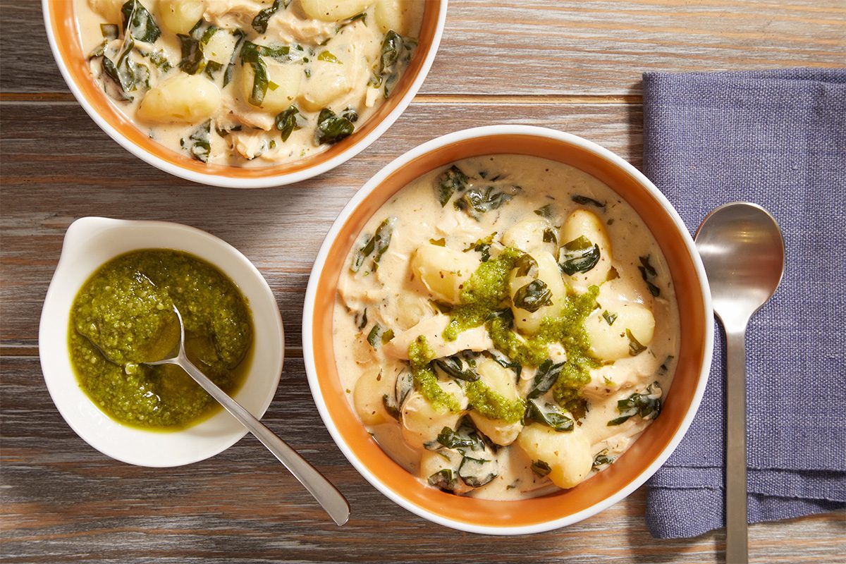 Two bowls of creamy gnocchi with spinach, topped with green pesto sauce, sit on a wooden surface beside a purple napkin, a spoon, and a small dish of extra pesto with a spoon.