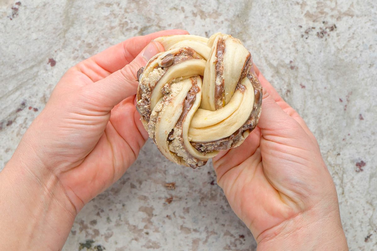 Two hands hold a twisted, unbaked pastry bun with visible layers of dough and chocolate filling over a light, speckled surface.