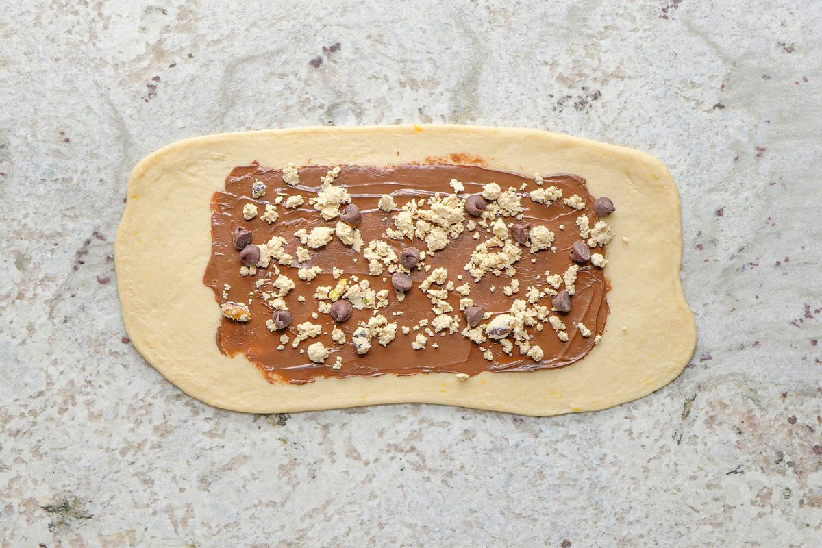 A rectangle of dough on a light countertop is spread with chocolate hazelnut spread and sprinkled with chopped nuts and chocolate chips.