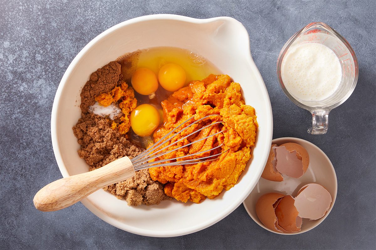 A bowl with brown sugar, pumpkin puree, three cracked eggs, and a whisk sits on a gray surface. Next to it are broken eggshells in a small bowl and a measuring cup filled with milk.