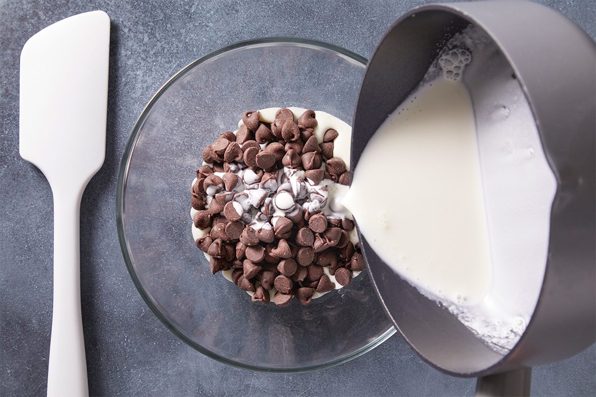 A glass bowl with chocolate chips and marshmallows is on a gray surface. Cream is being poured into the bowl from a gray measuring jug. A white spatula lies next to the bowl.