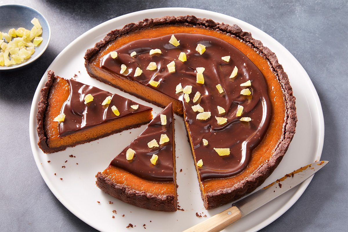 A chocolate and orange tart with a slice cut out, topped with glossy chocolate ganache and pieces of candied orange, sits on a white plate beside a knife.