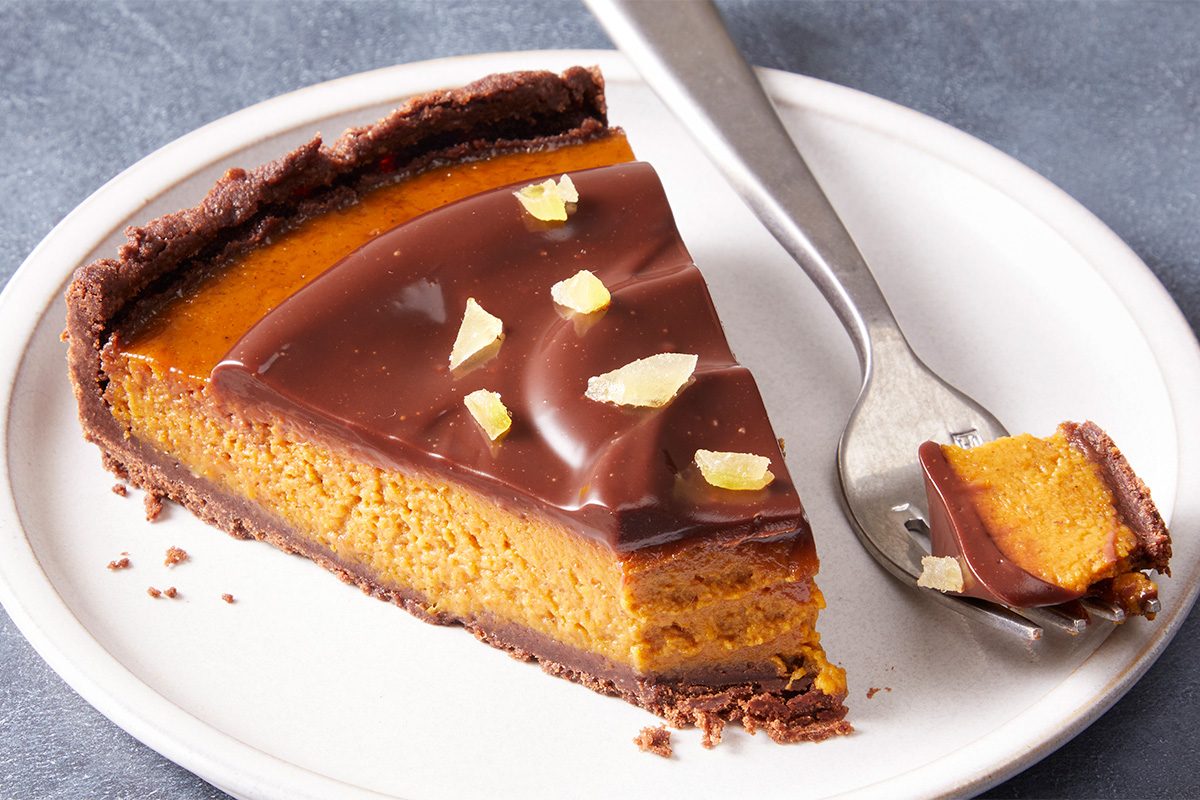 A slice of pumpkin pie with a chocolate topping and candied ginger pieces sits on a white plate. A fork holds a bite-sized piece of the pie.