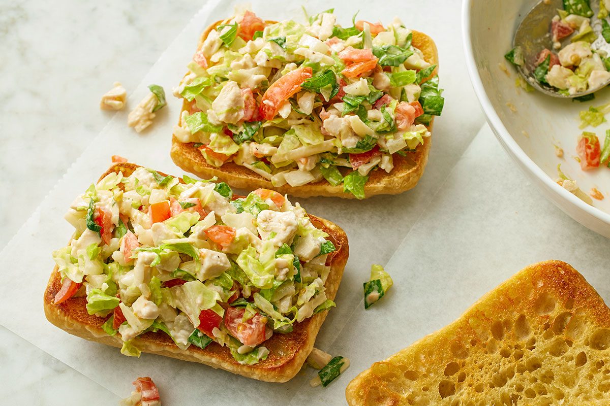 This is an overhead shot. Two open-faced sandwiches with chopped chicken salad; crisp lettuce; and juicy tomatoes on toasted ciabatta bread sit on a light background; nearby is a bowl of leftover salad with a spoon