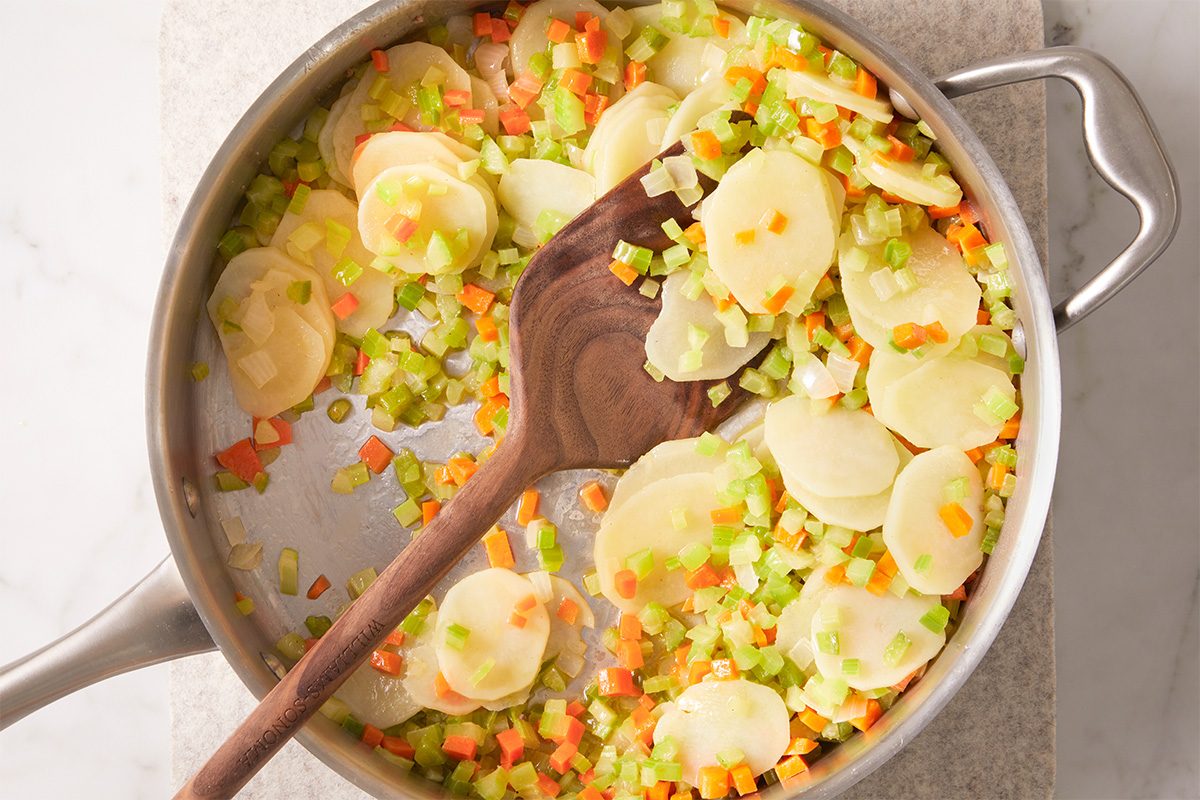 A stainless steel pan with sliced potatoes, diced celery, carrots, and onions being stirred with a wooden spoon on a light surface.