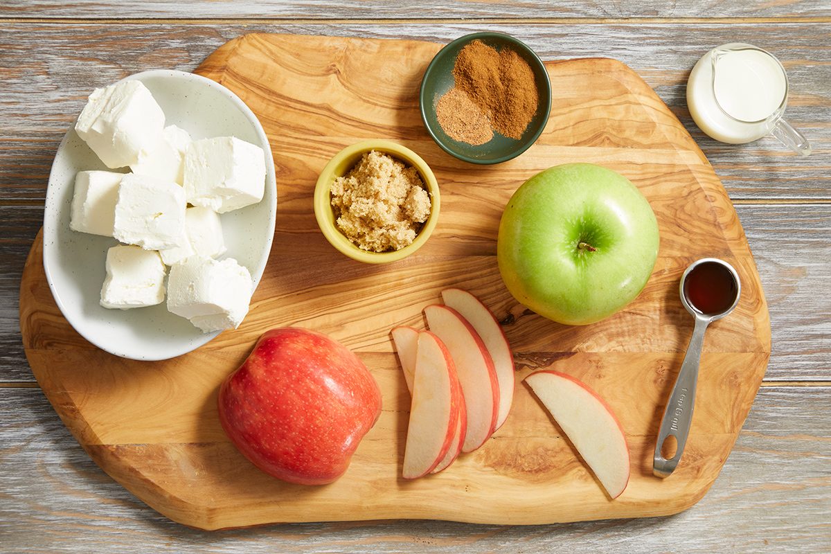 A wooden board with cubed cream cheese, green and red apples (whole and sliced), brown sugar, cinnamon, a small cup of milk, and a tablespoon of vanilla extract arranged neatly.