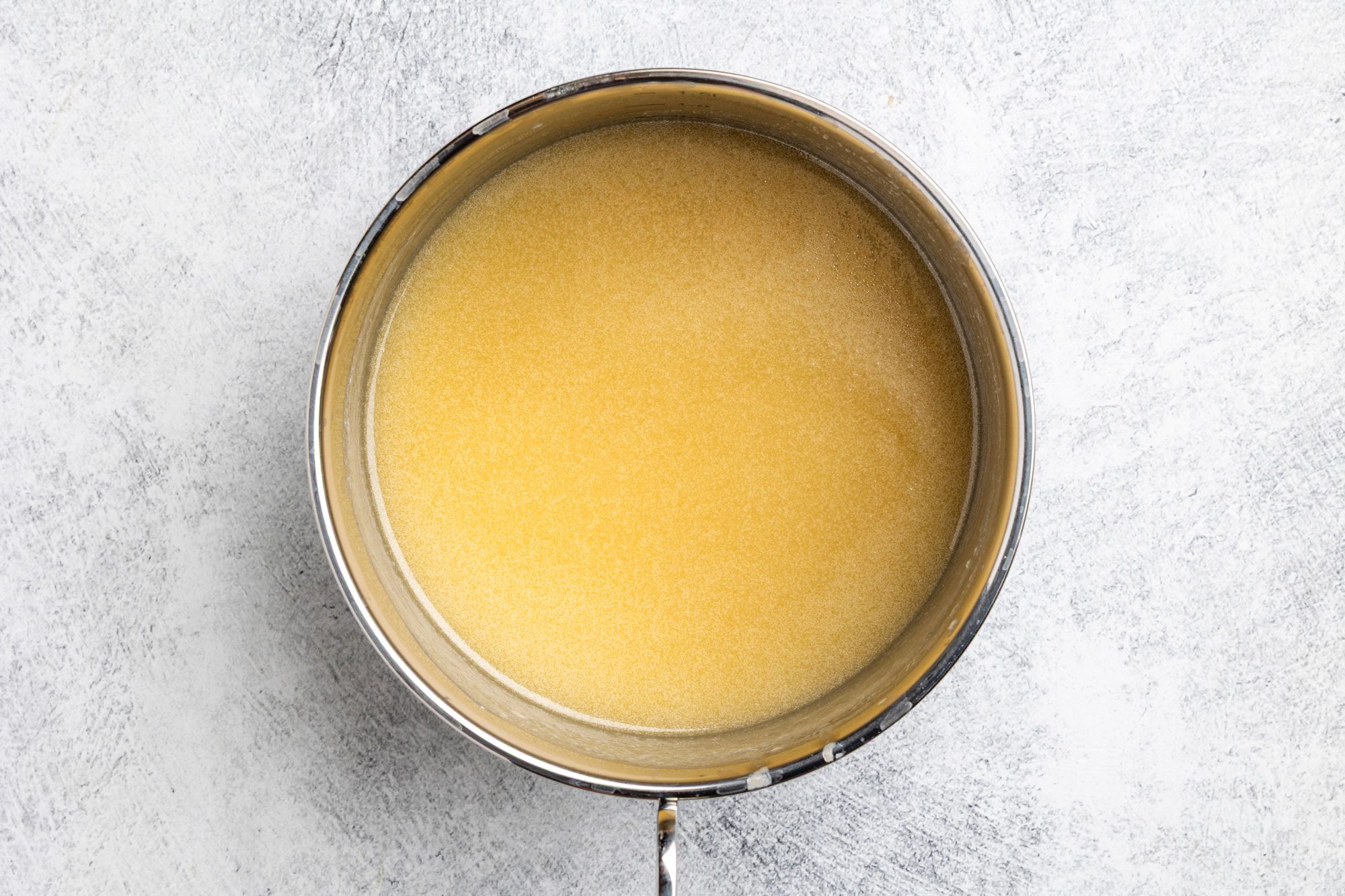 Overhead shot of a metal pot filled with light yellow liquid; possibly broth or stock; sitting on a white textured surface