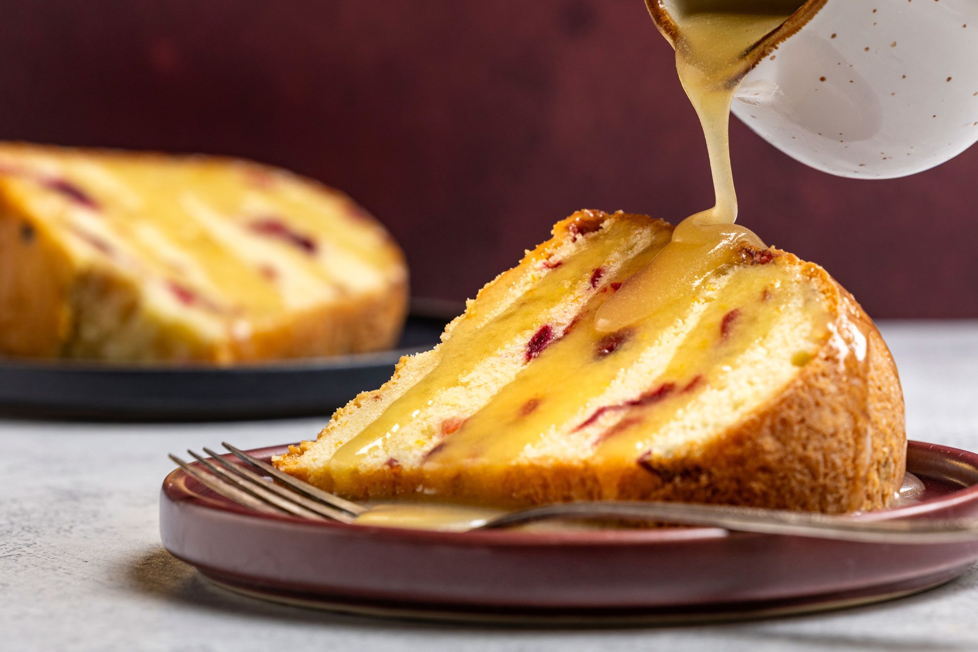 Close-up shot of a slice of Cranberry-Orange Pound Cake on a brown plate with a fork; as a rich golden glaze is being poured over the top; Another slice of cake is visible in the background; set on a dark plate against a deep red backdrop