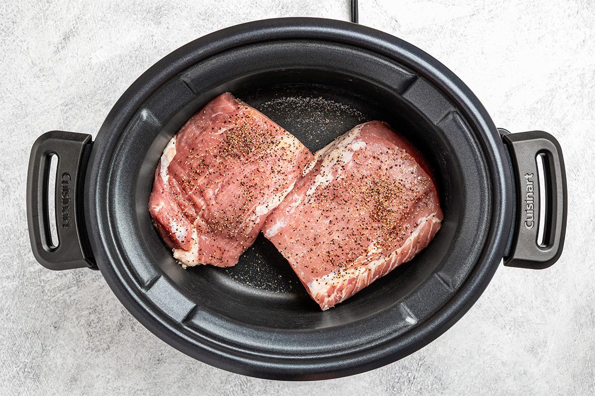 Two raw, seasoned pieces of pork sit side by side in a black slow cooker on a light gray countertop.