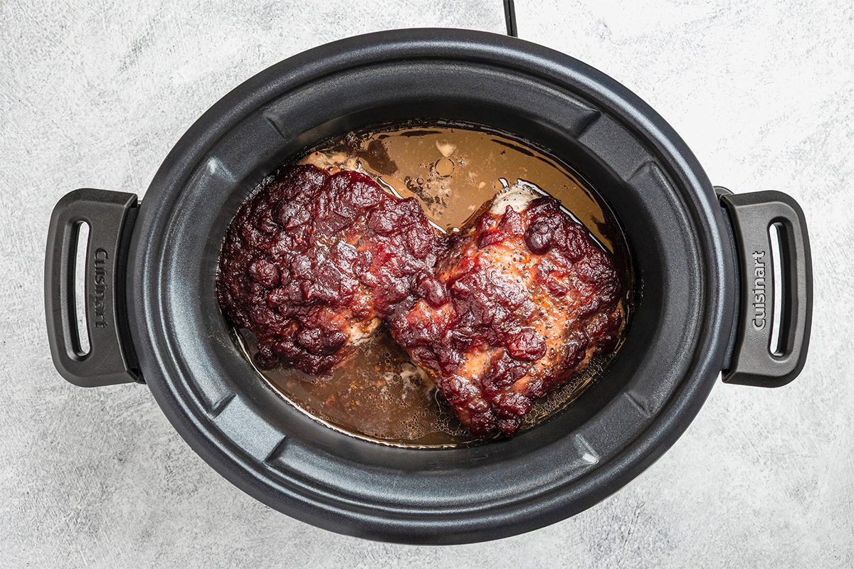 A slow cooker filled with two pieces of cooked meat covered in a dark, caramelized sauce, surrounded by juices, sitting on a light-colored countertop.