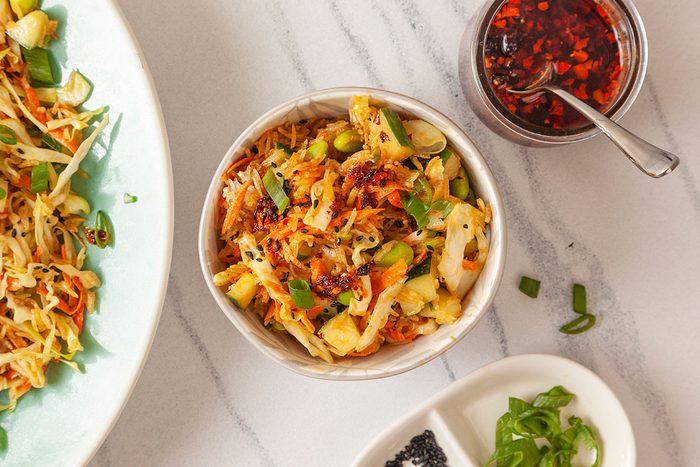 Close-up overhead shot of Crispy Rice Salad served in a small bowl, filled with shredded cabbage, carrots, edamame, and crispy rice, topped with chili flakes, sesame seeds, and green onions; Part of a larger plate of salad, a jar of chili oil with a spoon, and a small dish of green onions and black sesame seeds are visible nearby on the marble surface;
