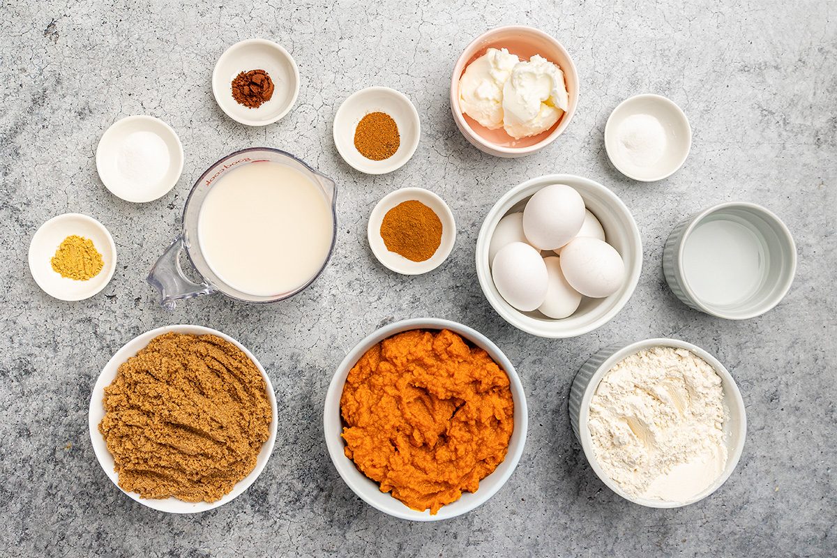 Various baking ingredients in bowls on a gray surface, including brown sugar, pumpkin puree, flour, eggs, milk, spices, baking powder, baking soda, shortening, and water, all arranged neatly for a recipe.