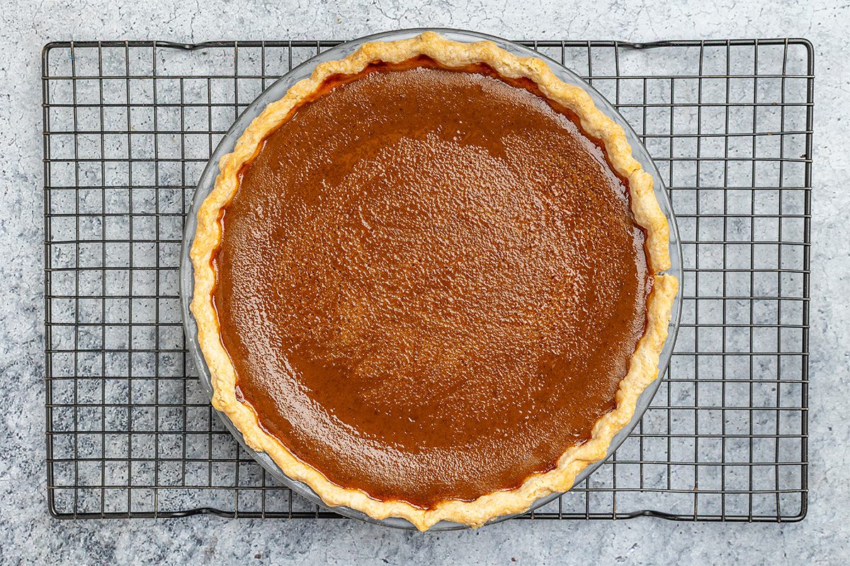A whole pumpkin pie with a golden, flaky crust sits on a round metal cooling rack, viewed from above against a gray textured background.