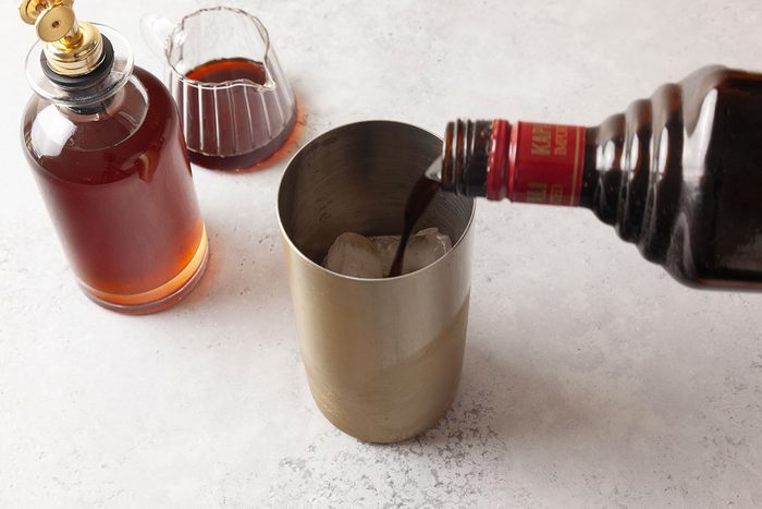 A bottle pours a dark liquid over ice in a metal cocktail shaker. Nearby are a glass decanter and a small glass with similar dark liquid, all on a light-colored surface.