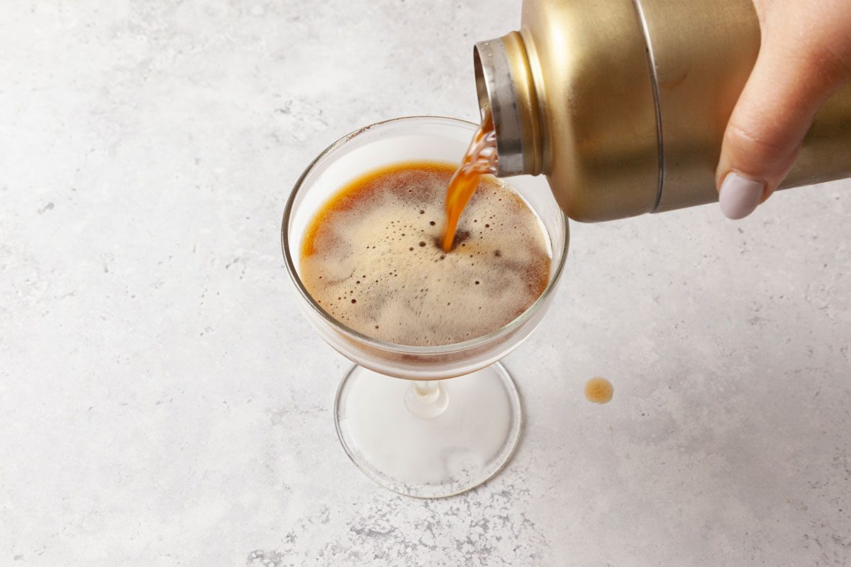 A close-up of a hand pouring a dark, foamy espresso martini from a gold cocktail shaker into a glass on a light, textured surface.