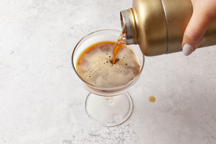 A close-up of a hand pouring a dark, foamy espresso martini from a gold cocktail shaker into a glass on a light, textured surface.