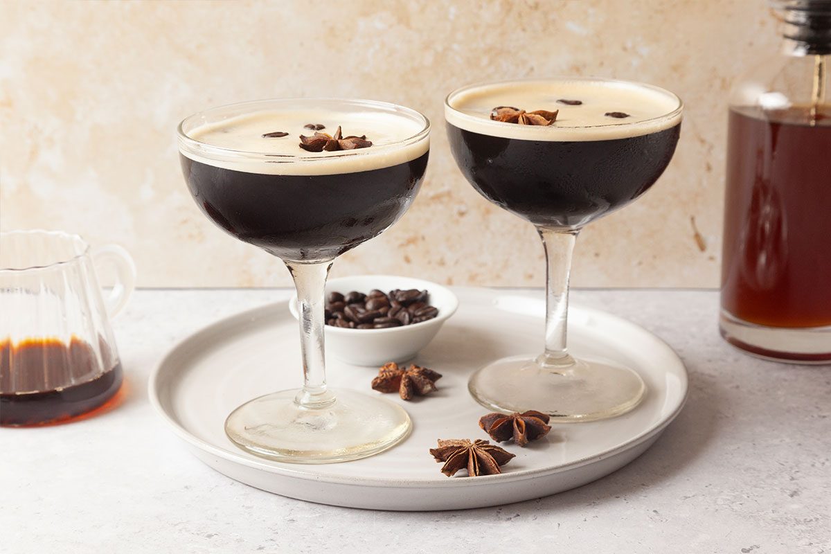 Two espresso martinis in coupe glasses on a white tray, garnished with coffee beans and star anise. A small bowl of coffee beans and a glass container with liquid are also on the tray, with a bottle in the background.