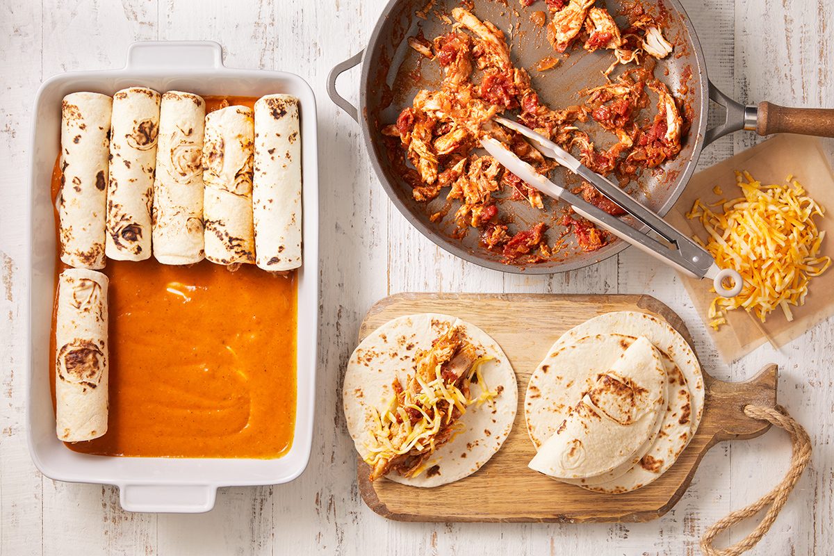 A baking dish with sauce and rolled tortillas, a skillet with chicken filling, shredded cheese on a board, and two tortillas being assembled on a wooden board. Ingredients are spread on a white wooden surface.