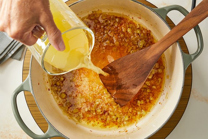 A hand pours broth from a glass measuring cup into a pot with sautéed onions, stirring with a wooden spatula.