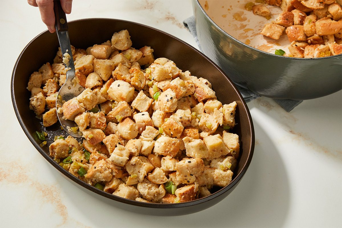 A hand uses a spatula to serve bread stuffing from an oval baking dish. The stuffing is golden brown with visible herbs and vegetables. A pot with more stuffing sits nearby on a white countertop.