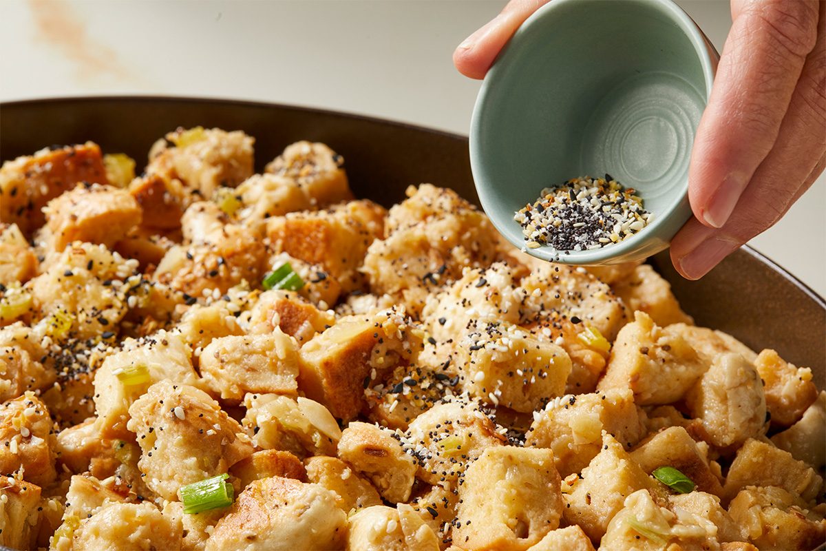 A hand sprinkles a blend of seeds and spices from a small bowl onto a pan of cubed bread stuffing with chopped green onions.