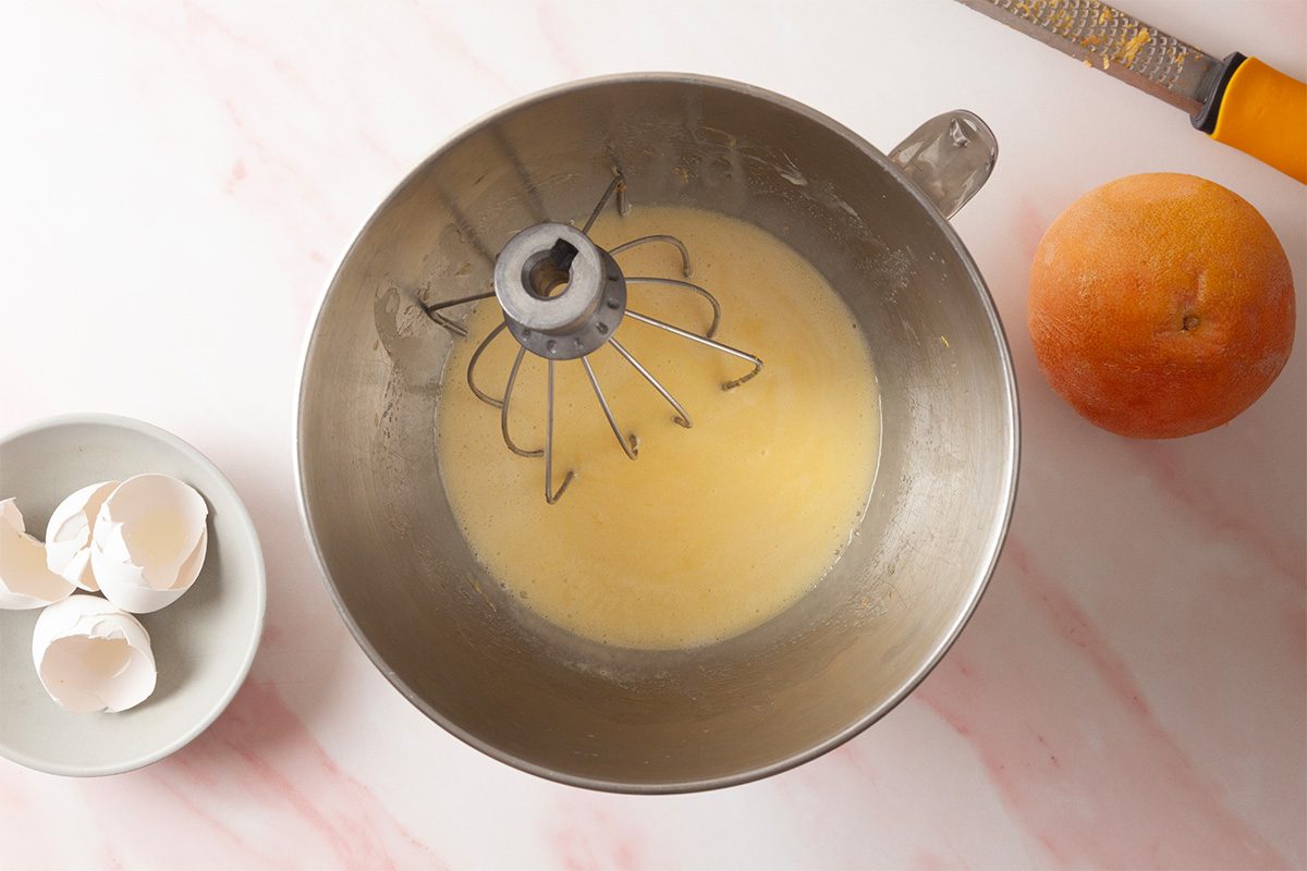 A metal mixing bowl with batter and a whisk attachment sits on a marble surface. Nearby are a zester with an orange handle, a whole orange, and a small bowl containing cracked eggshells.