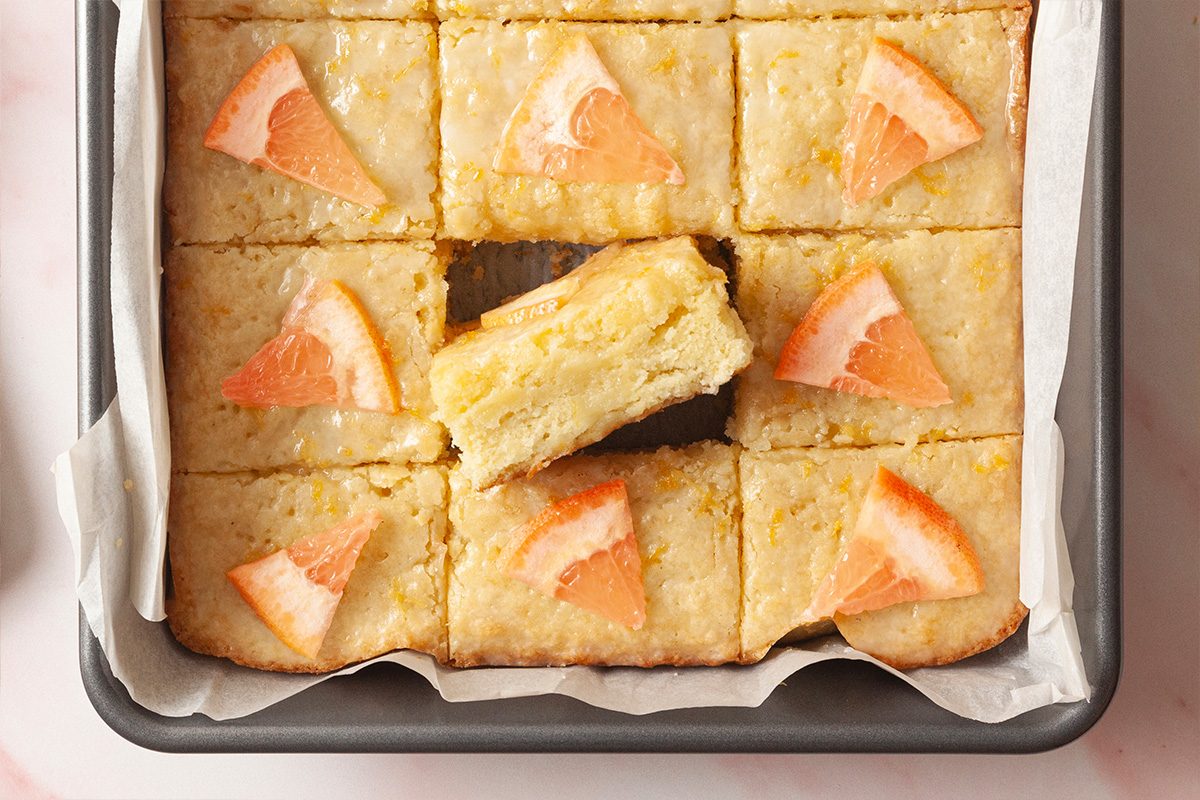 A pan of grapefruit-topped blondies cut into 12 squares, with one piece stacked on top of another, showing the moist interior. The bars are lightly golden with small grapefruit segments on top.