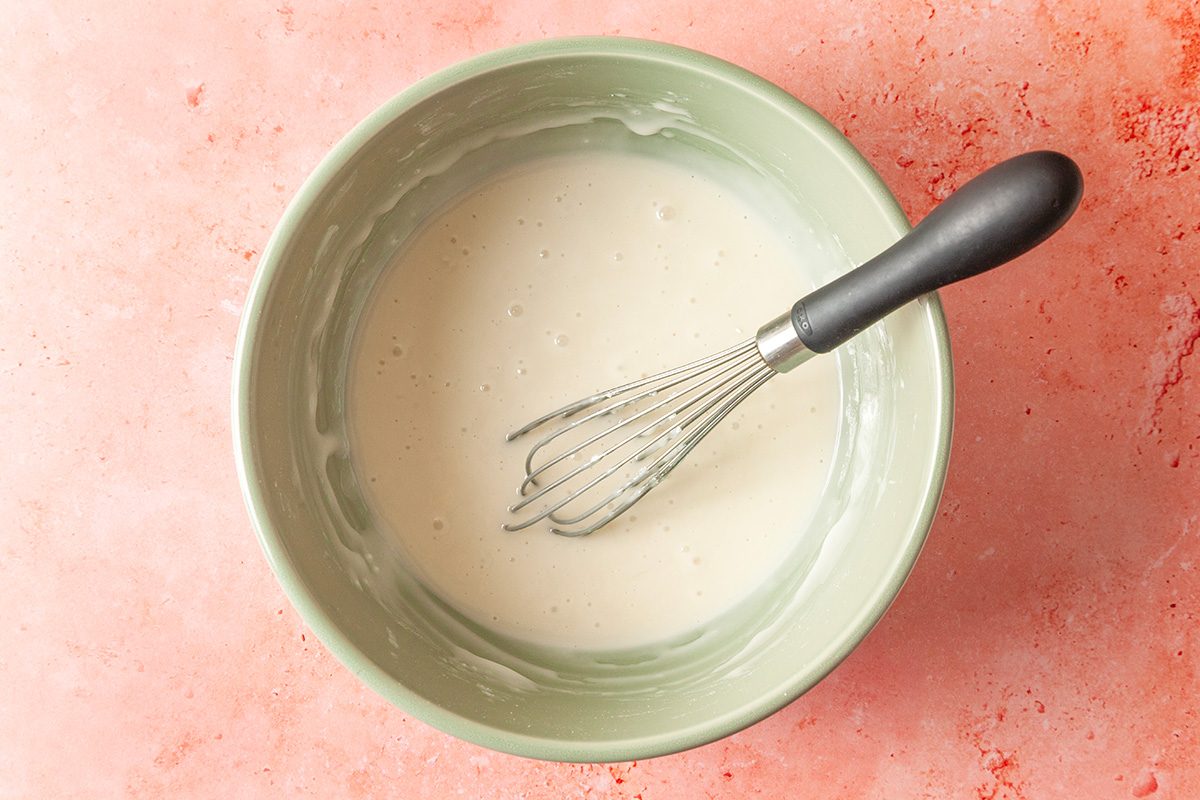 Overhead shot of a green bowl filled with smooth white batter, being whisked with a black-handled metal whisk, set on a light pink countertop