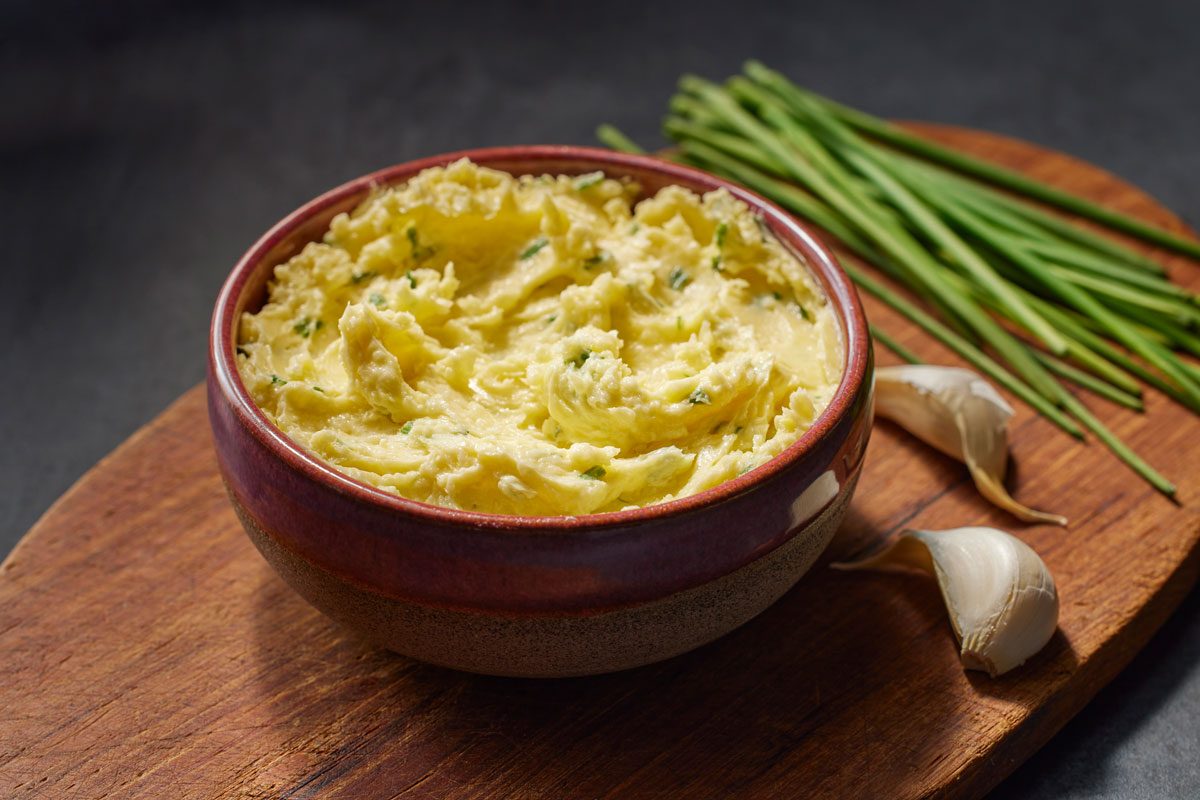 3/4th shot of Garlic Chive Butter Creamy mashed potatoes topped with chives in a bowl on a wooden board, fresh chives and garlic beside it