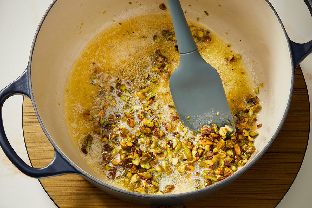 A saucepan with melted butter and chopped pistachios being stirred with a blue spatula, sitting on a wooden surface.