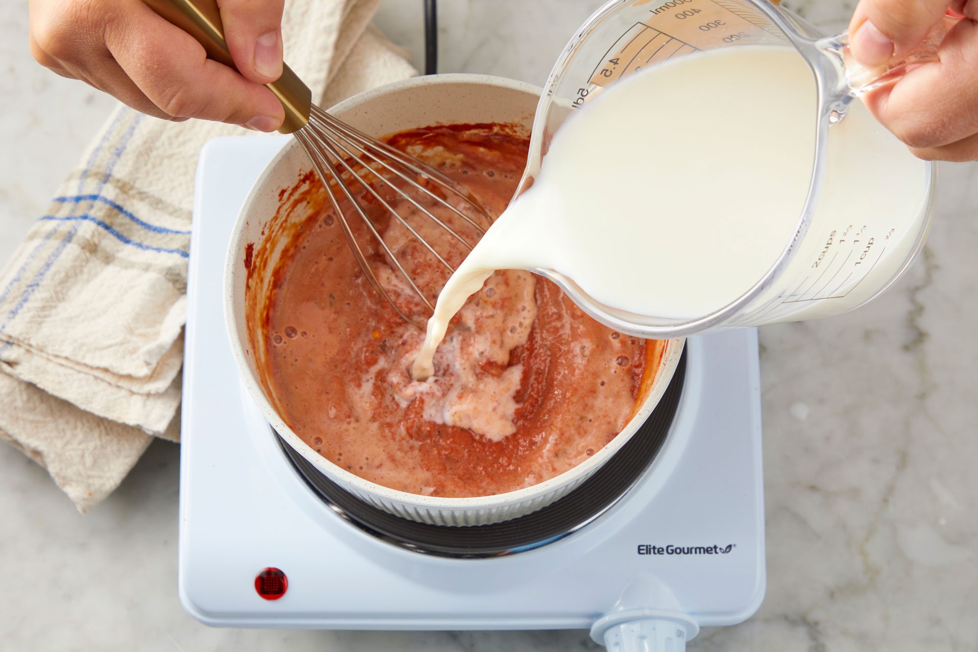 Overhead shot of a small saucepan combine tomato paste, garlic, salt, pepper and remaining 1/2 teaspoon basil; cook and stir over medium heat for 1 minute; gradually whisk in milk; bring to a boil; reduce heat; simmer uncovered for 4-5 minutes or until thickened; stirring frequently; Remove from heat; a napkin nearby; all set on a marble surface;