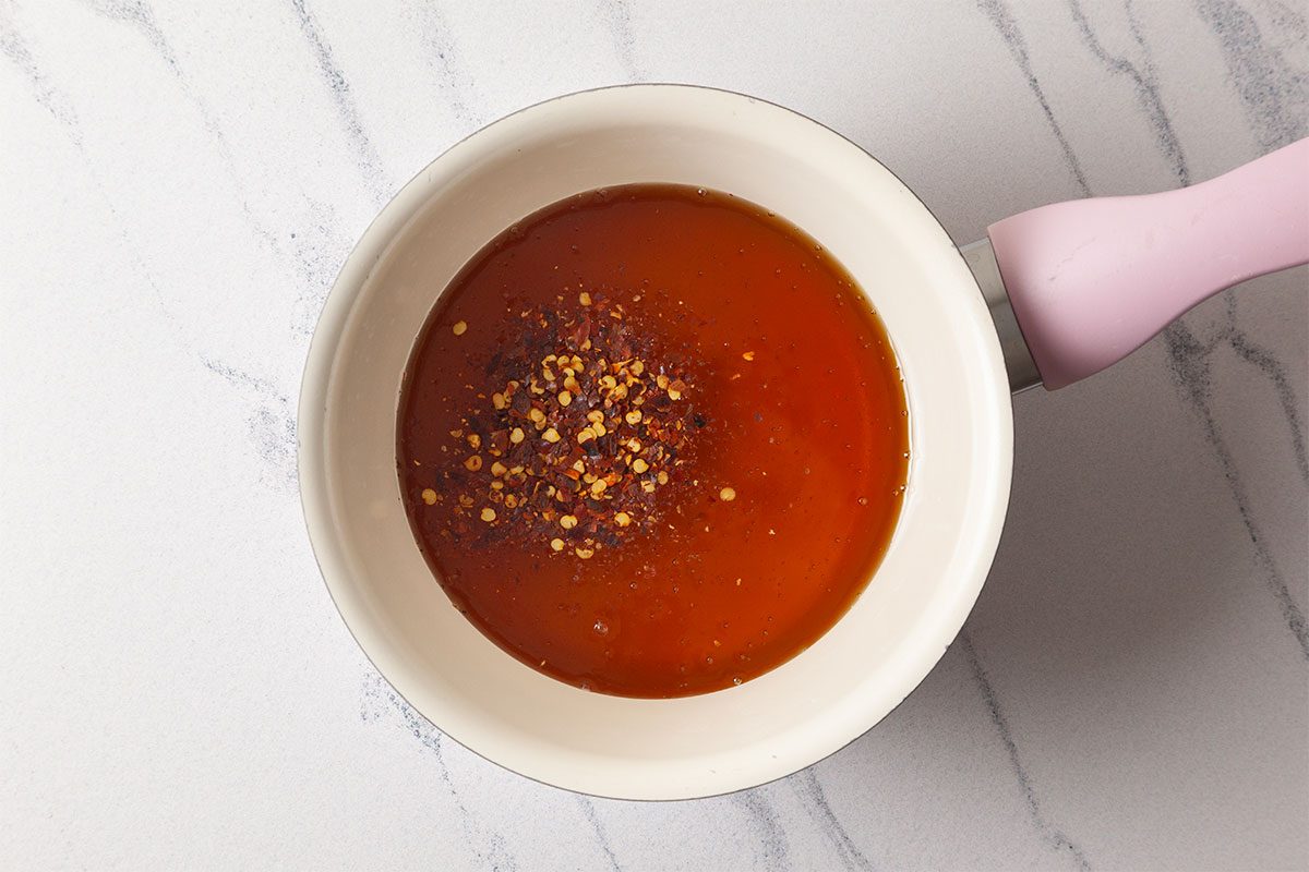 A white saucepan with a pink handle contains a reddish-orange sauce topped with crushed red pepper flakes, sitting on a white marble surface.
