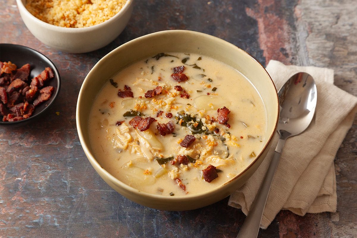 A bowl of creamy soup topped with crumbled bacon and shredded cheese sits on a napkin with a spoon beside it; a small bowl of cooked grains and a dish of more bacon pieces are in the background.