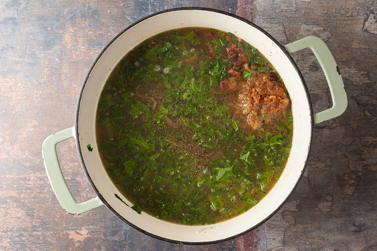 A large pot filled with broth, fresh chopped herbs, and pieces of meat sits on a textured surface.