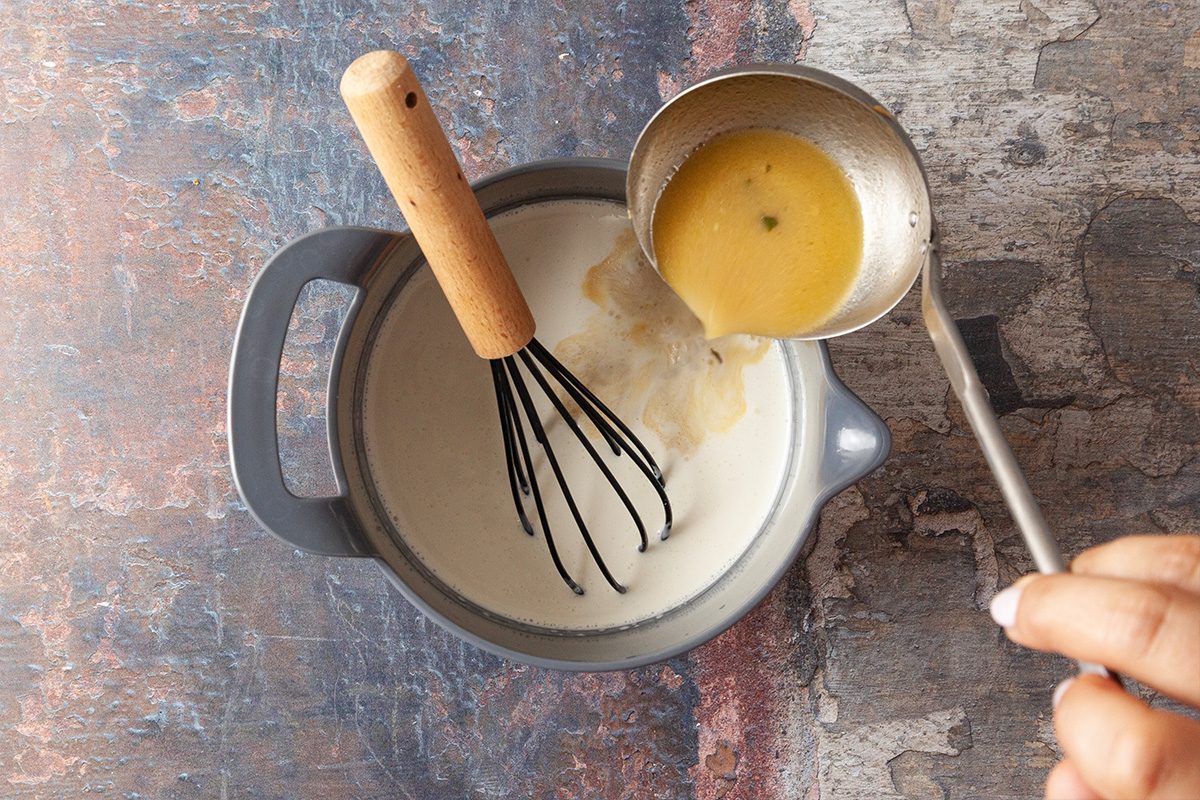 A hand pours a yellow liquid from a ladle into a pot of white liquid with a whisk inside, on a textured surface.