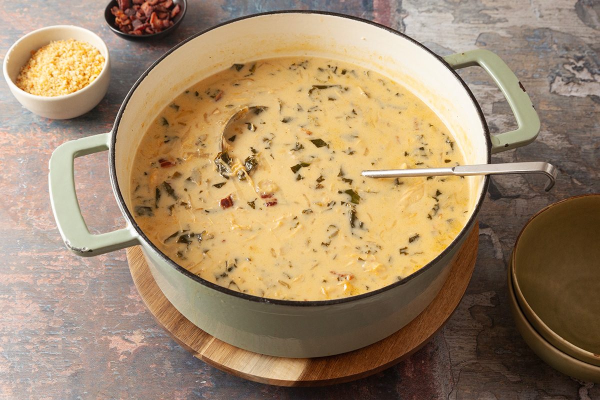 A large pot of creamy soup with leafy greens, bacon pieces, and a bay leaf, sits on a wooden trivet with a ladle inside. Nearby are bowls of crispy bacon bits and shredded cheese.
