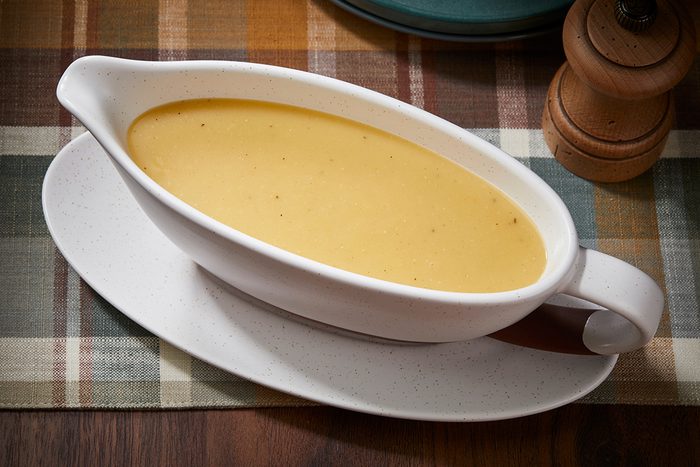 A white gravy boat filled with creamy yellow sauce sits on a matching oval plate atop a plaid tablecloth, with a pepper grinder in the background.