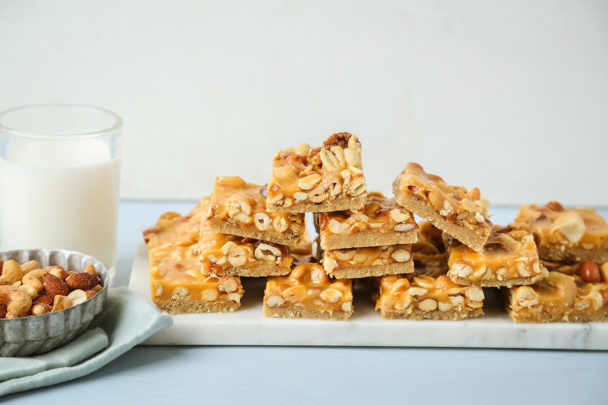 A stack of nut-topped cookie bars sits on a marble tray next to a glass of milk and a small bowl of mixed nuts, all placed on a light blue surface.