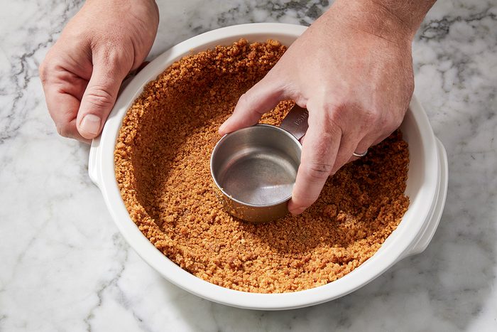 A person presses a metal measuring cup into a pie dish filled with a crumbly graham cracker crust, shaping it evenly against the sides and bottom on a marble countertop.