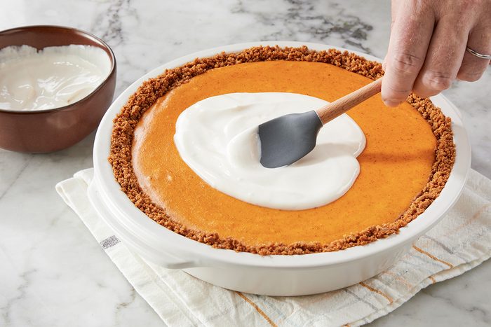A hand uses a spatula to spread a layer of whipped cream on top of a pumpkin pie with a crumb crust. A bowl of extra whipped cream sits nearby on a marble surface.