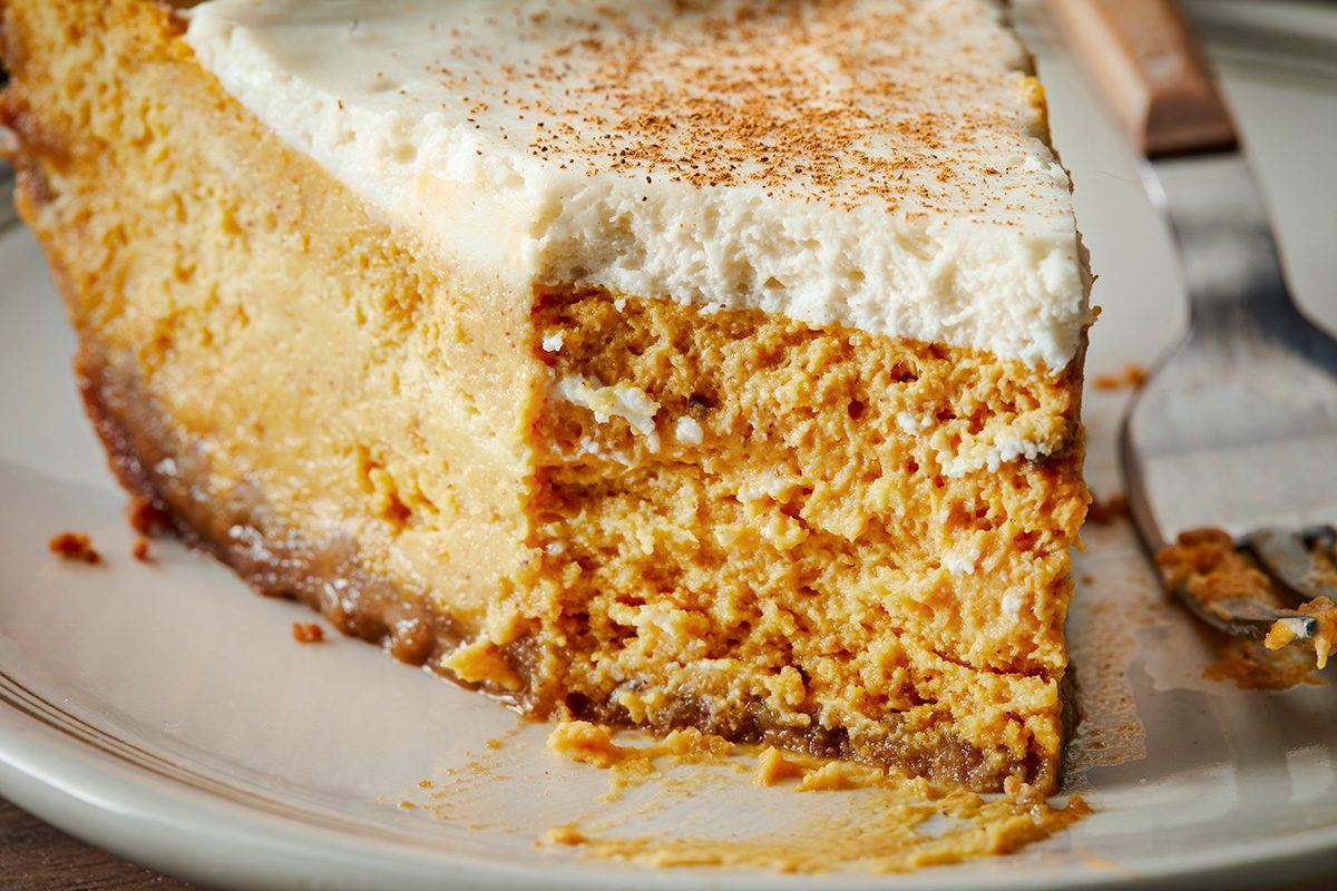 A close-up of a slice of pumpkin cheesecake topped with a layer of whipped cream and a sprinkle of cinnamon, showing its creamy texture and crumbly crust on a white plate.