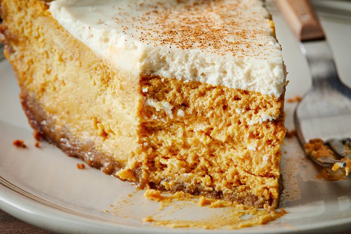 A close-up of a slice of pumpkin cheesecake topped with a layer of whipped cream and a sprinkle of cinnamon, showing its creamy texture and crumbly crust on a white plate.