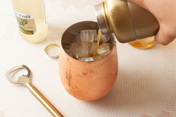 A hand pours a drink from a gold shaker into a copper mug filled with ice cubes. A bottle, bottle cap, and gold bottle opener are on a white surface nearby.