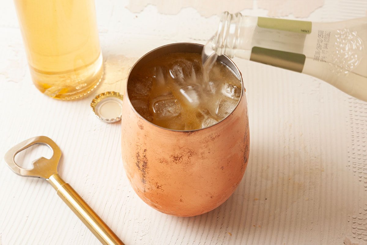 A bottle pours a clear liquid over ice into a copper mug on a white surface, next to a bottle opener, a bottle cap, and a glass bottle with a yellow beverage.