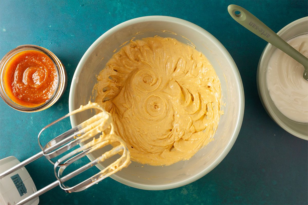A mixing bowl filled with creamy batter, an electric hand mixer with beaters resting inside, a small bowl of orange puree, and another bowl with white mixture on a teal surface.