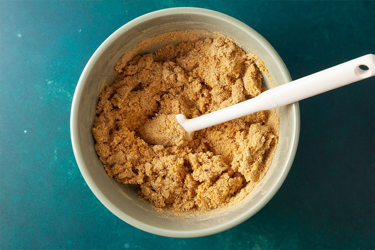 A mixing bowl filled with a crumbly brown sugar and flour mixture, with a white spoon resting inside, sits on a teal surface.