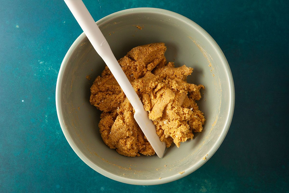 A bowl of crumbly, light brown dough with a white spatula resting inside, viewed from above on a teal surface.